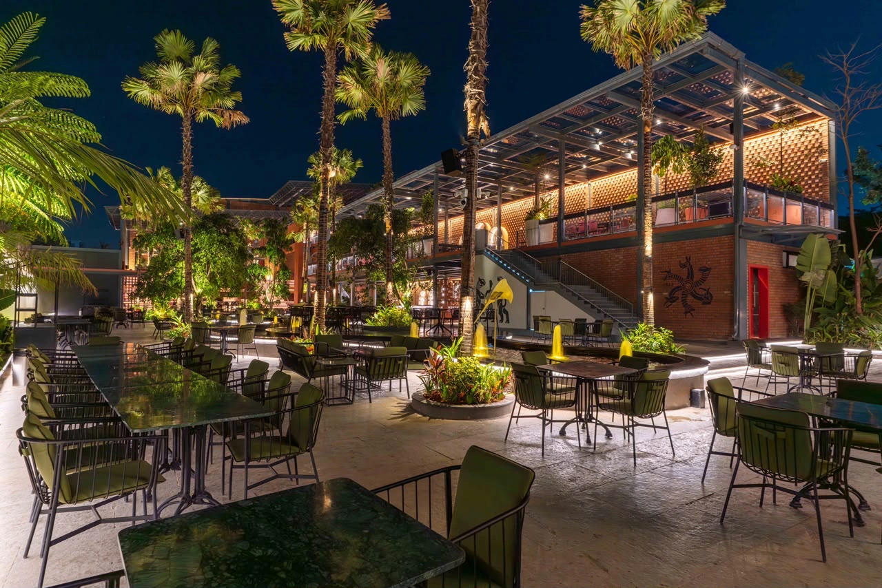 The ICB courtyard at night: palms lit from below, green chairs at marble tables, and the brick bar building with its painted pictogram and steel canopy