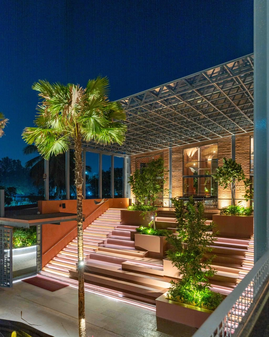 Lit stepped seating with planters rising under the steel space frame, a palm in front and the glasshouse behind